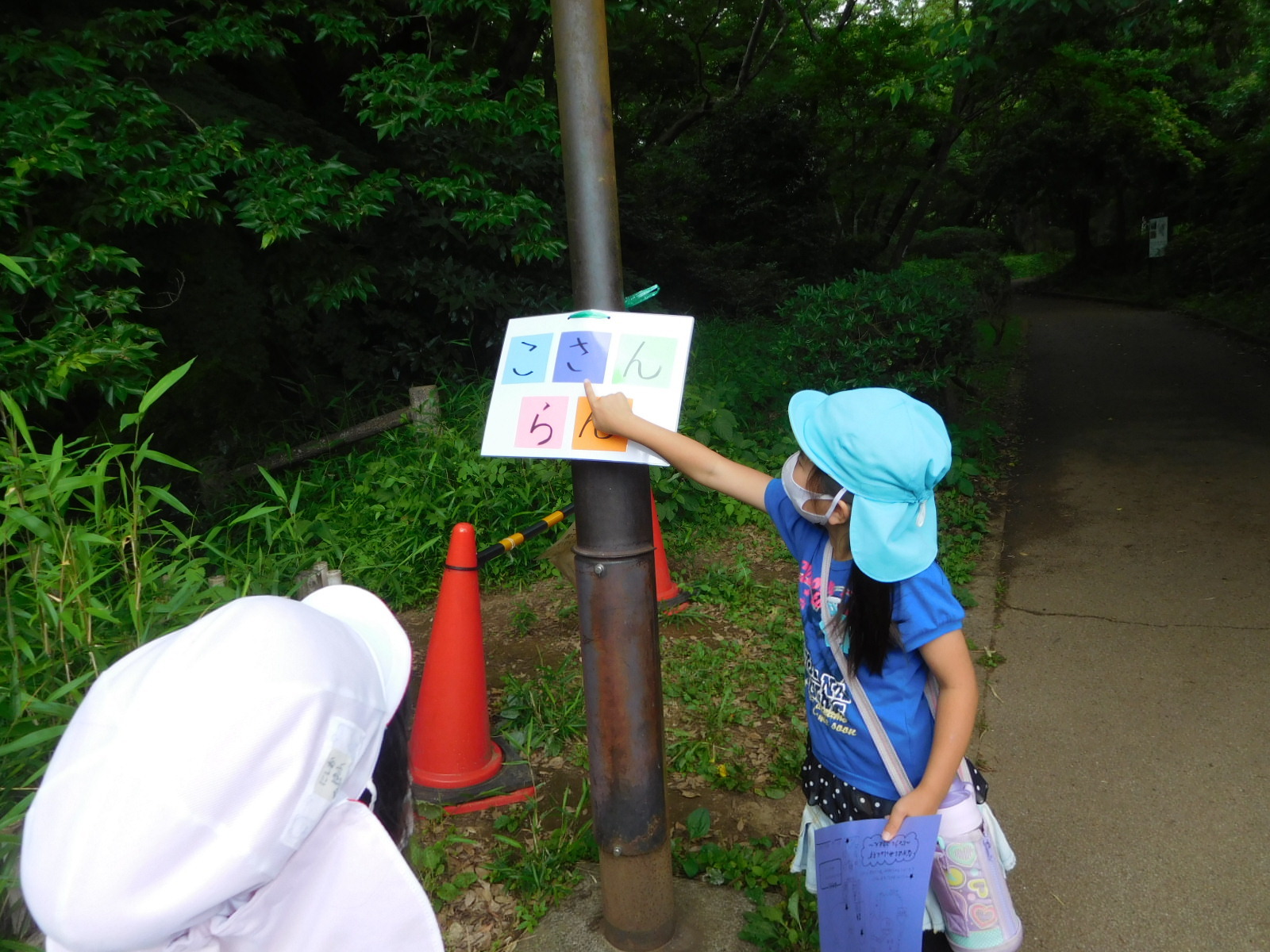 公園内の鉄の柱のようなものにつけられた5文字の文字が書かれたカードを指さす女の子の園児の写真