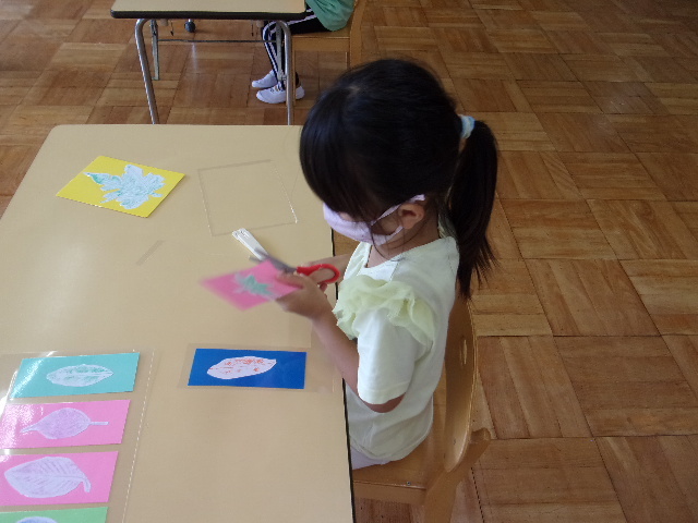 葉っぱのスタンプが貼られた色紙が、ラミネートされ、女の子の園児が、はさみで切っている写真