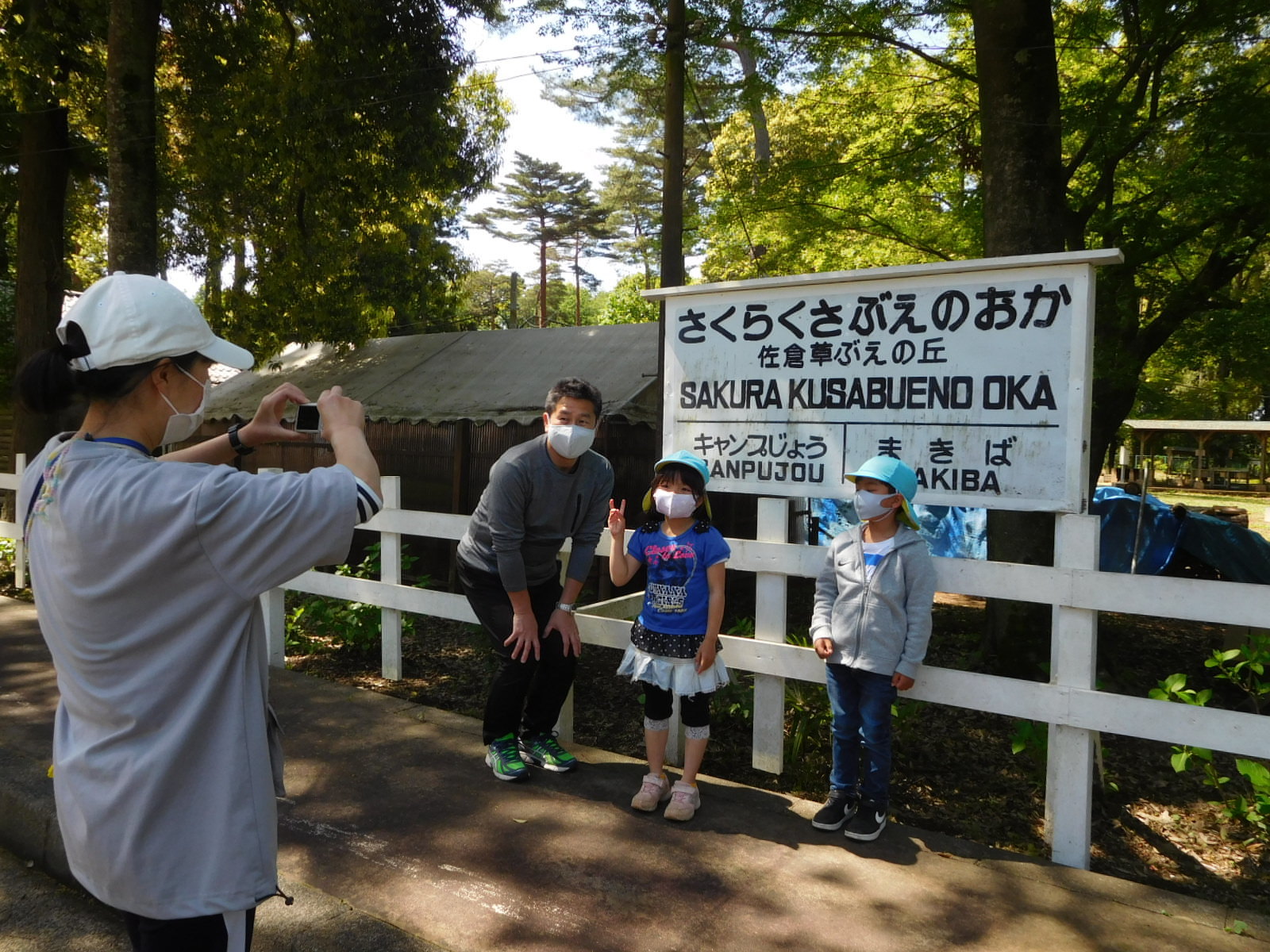 「さくらくさぶえのおか」と書かれた駅名の看板の前で記念撮影をする子供たちと先生の写真