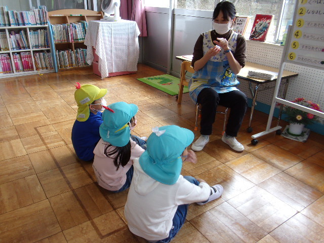 床に体育座りをしている3人の園児の前で、椅子に座った先生が話をしている写真