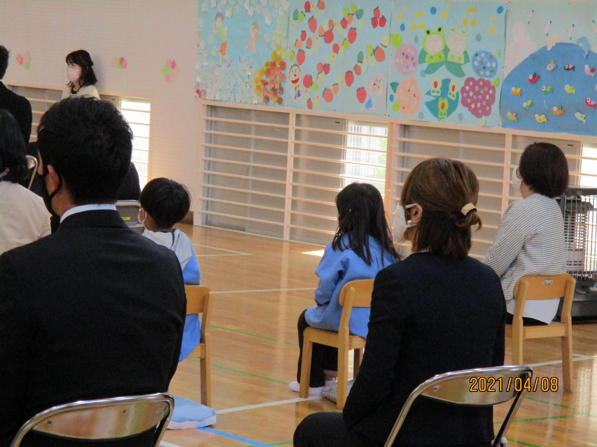 入学式の参加者が着席している中に座っている園児2名の後ろ姿の写真
