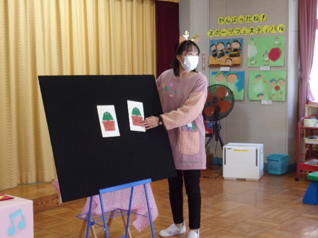 木のイラスト2枚が貼ってある黒いパネルの横に、トナカイのカチューシャを付けた先生が立っている写真