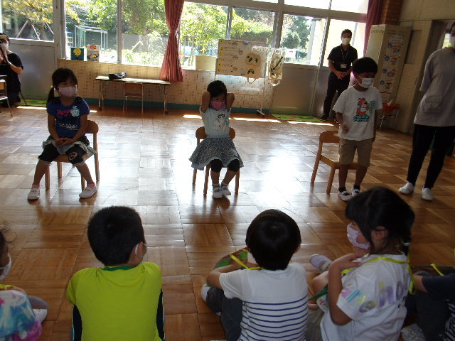 並んで座っている児童の前で、2名の園児は椅子に座り、男の子の園児は立っている写真