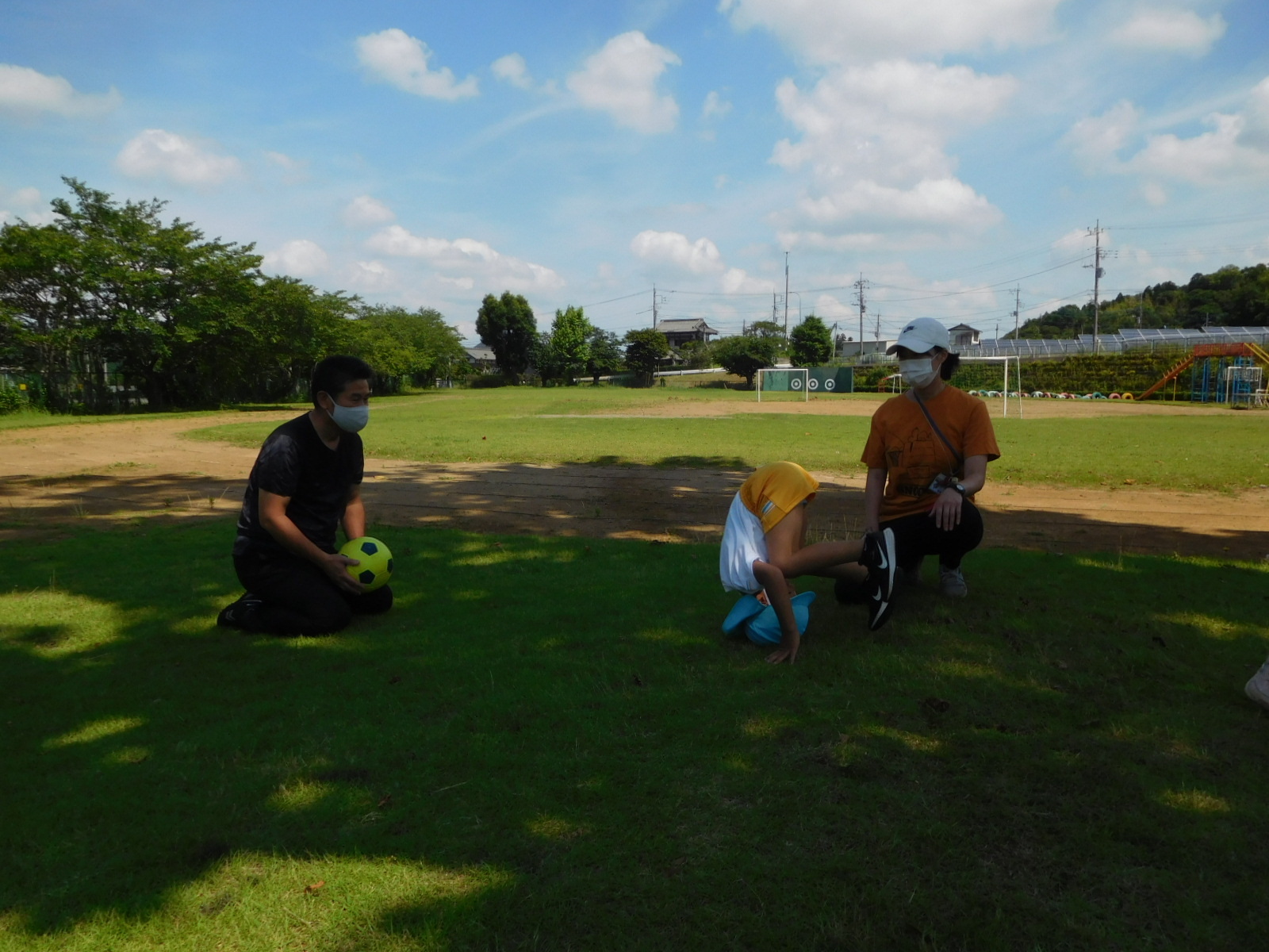 木陰の芝生の上で、前回りをする園児と、ボールを持って座っている園長先生の写真
