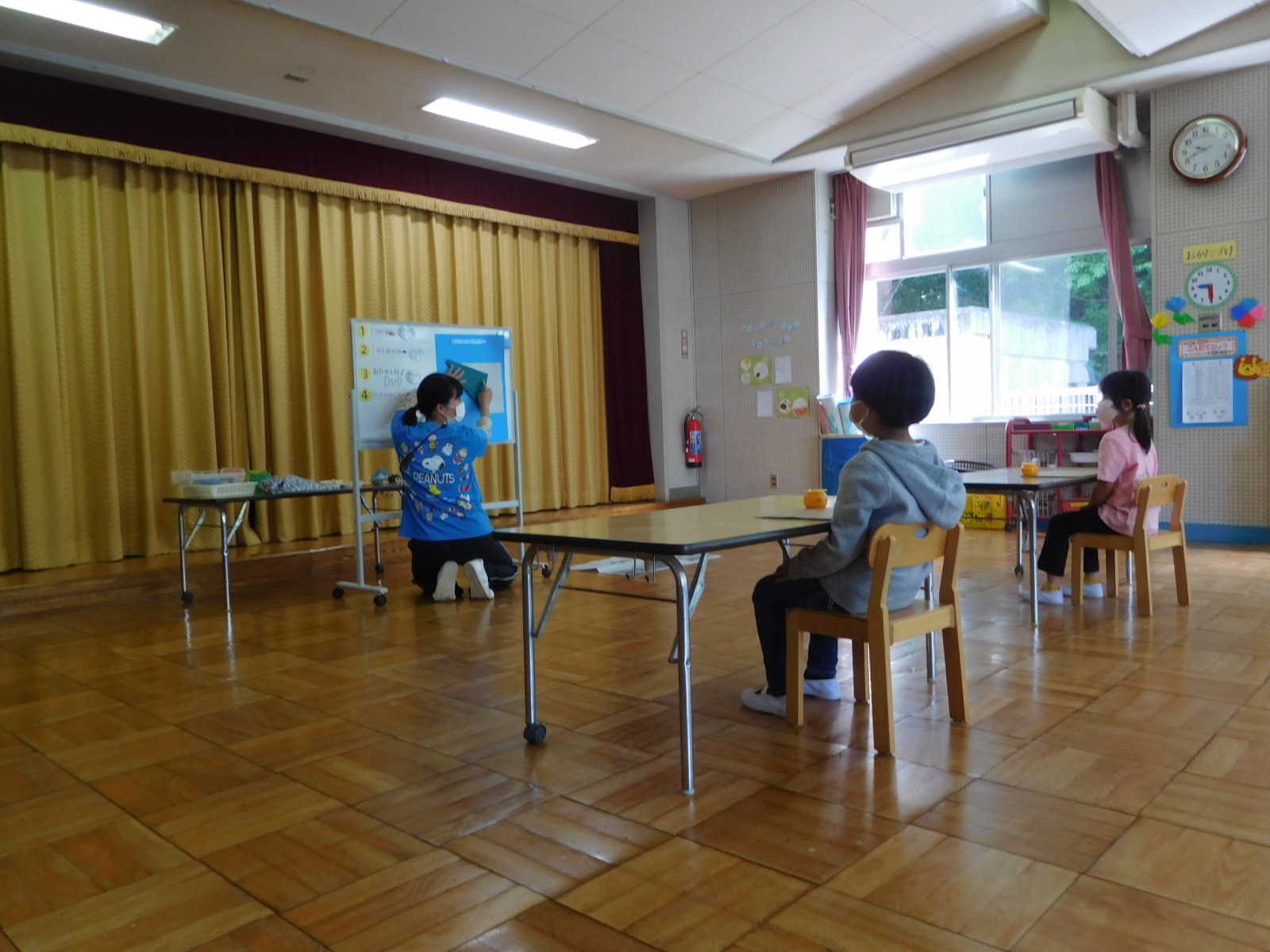着席している男の子と女の子の園児の前で、先生がホワイトボードの前で織り方を教えている写真