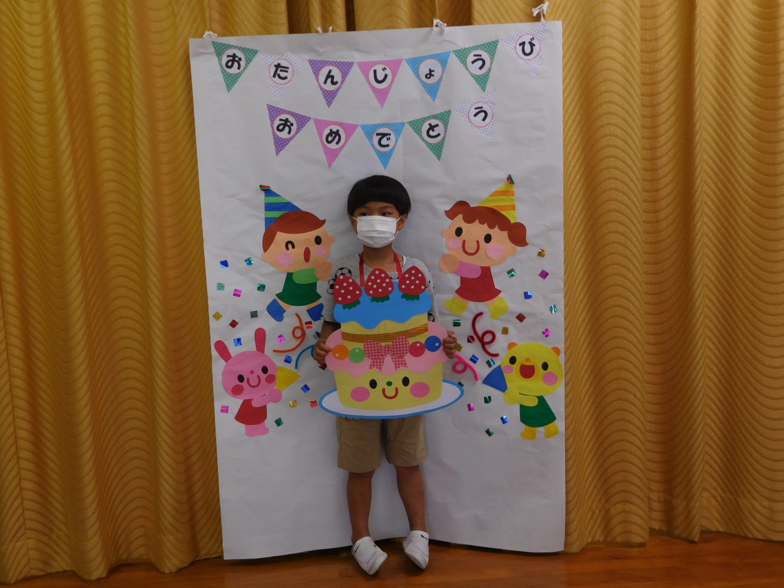 お誕生日パネルの前に立ち、ケーキの絵を持っている男の子の園児の写真