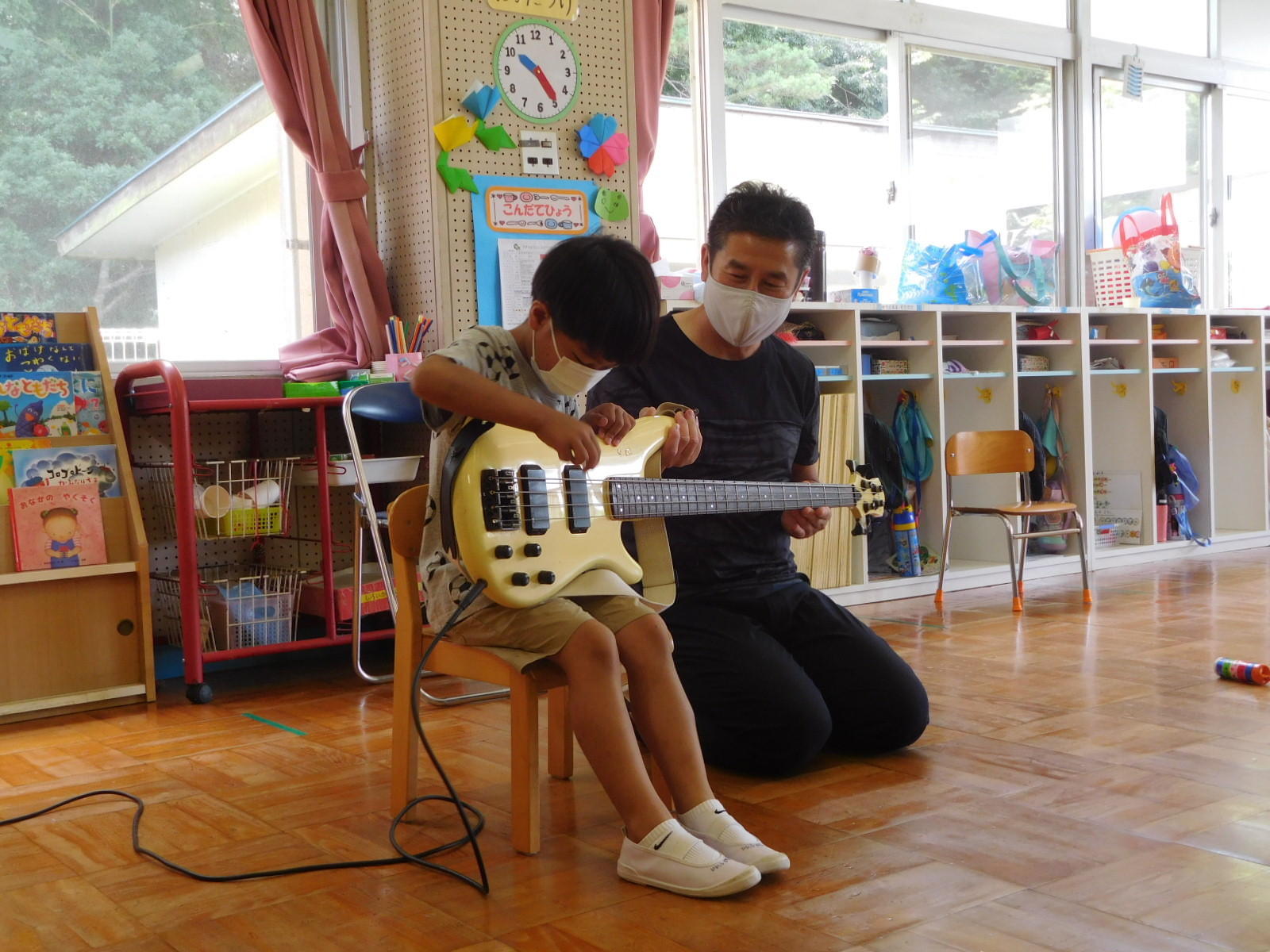 男の子の園児が、園長先生に支えられながらベースを弾いている写真
