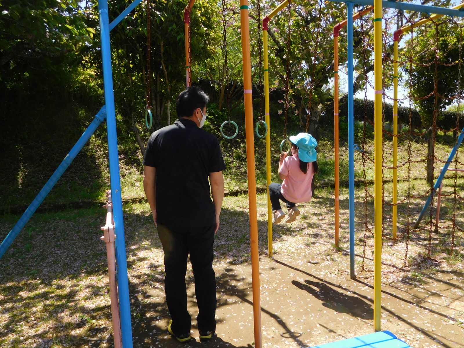 吊り輪ジムにぶらさがる水色のカラーキャップを被った女の子の園児と、その横に立つ男性の写真