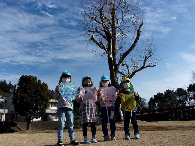 すずかけの木の前で、手作りの凧を持って並んでいる園児4名の写真