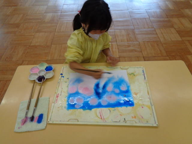 黄色いスモックを着た女の子の園児が、画用紙に絵の具で色を塗っている写真