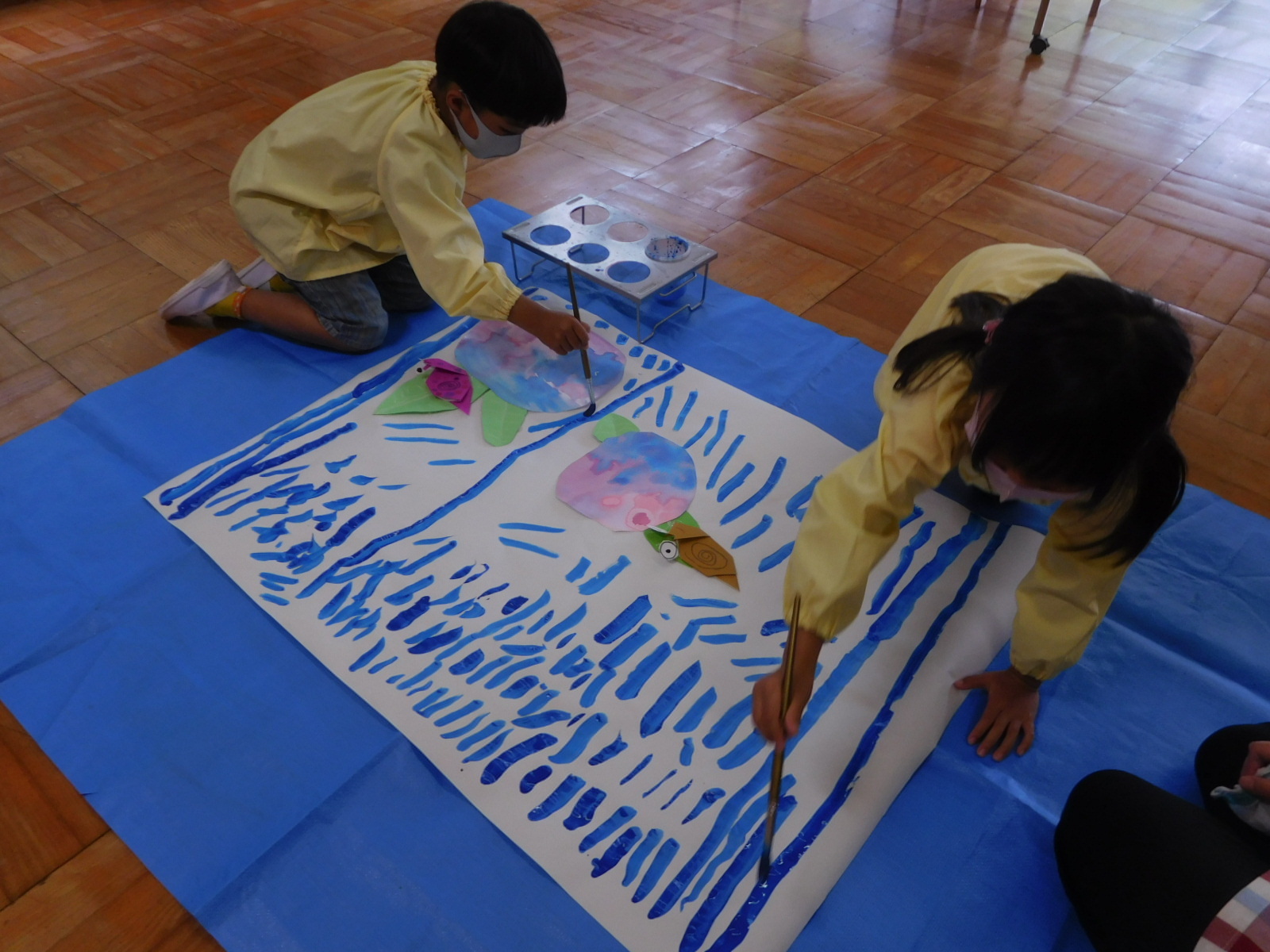 ブルーシートの上の大きな模造紙に手作りのアジサイとカタツムリが貼られ、2名の園児が青い絵の具で雨を降らせている写真