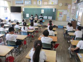 教室内で3年生の3人が歌詞を読んでいる様子を椅子に座ってみている1年生達の写真