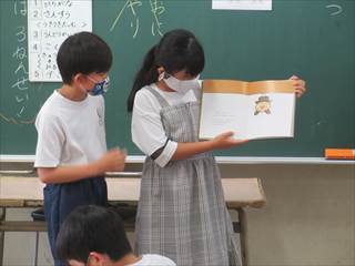 教室内で4年生の女子生徒が本を開いていて男子生徒がその本の朗読をしている様子の写真