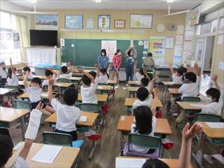 教室内で5年生の3人が黒板の前で並んでいて椅子に座ってみている1年生達が手を上げている様子の写真