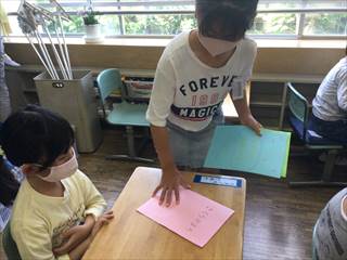 教室で6年生の生徒が桃色の歌詞カードを1年生の机に配っている写真