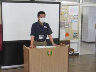 教壇に両手を付いている紺色のネルシャツ姿でマスクを付けている校長先生の写真