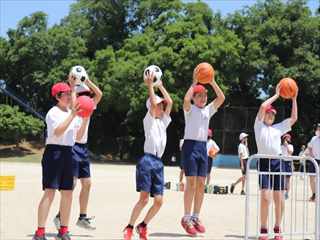 校庭で体操着姿の6年生がバスケットボールやサッカーボールを上に掲げている写真