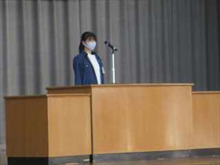 後期終業式で壇上に立ってマイクに話し掛けている紺色の服を着た女の子の写真
