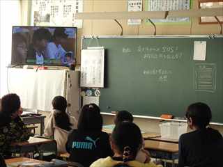 教室で自分の机に座りながら左側に設置されたモニターの映像を見ている子どもたちの写真