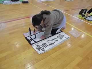 体育館の床に半紙を広げ、集中した様子で書き初めをしている女の子の写真