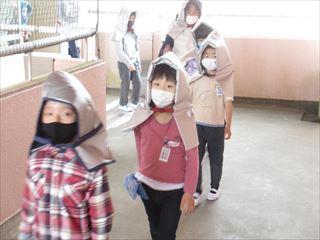 避難訓練で銀色の防災頭巾とマスクを付けて廊下を移動している生徒たちと引率している教師の写真