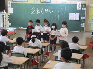 教室内で2年生の5人が問題を読んでいる様子を椅子に座ってみている1年生達の写真