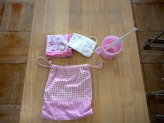 ピンクの柄のナプキンと白いマスクと歯ブラシ、コップとそれらを入れるピンク色のチェック柄の給食袋の写真