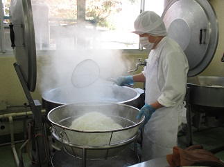 白衣の帽子とマスク、アイお手袋をした作業員が大きな鍋から食材を移している様子