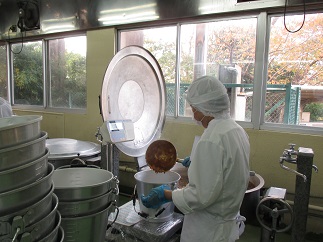 白衣の帽子とマスク、青い手袋をした作業員ができた食材を別の容器に移している様子