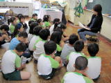 多数の児童が、教室内前方で体育座りをし、椅子に座ったボランティアの方の読み聞かせを聞いている写真