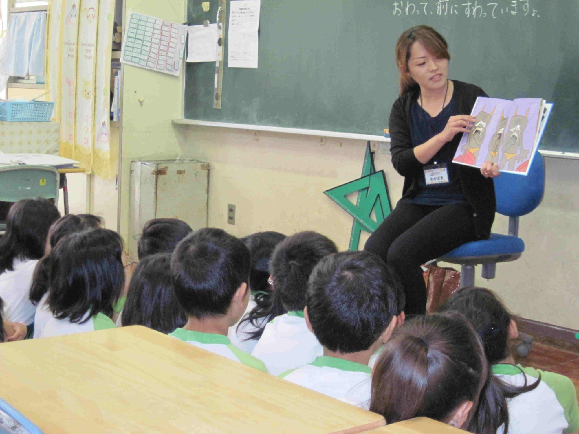 椅子に座りながら読み聞かせをするボランティアの方と、多数の児童が、黒板前に集まり、読み聞かせを聞いている写真