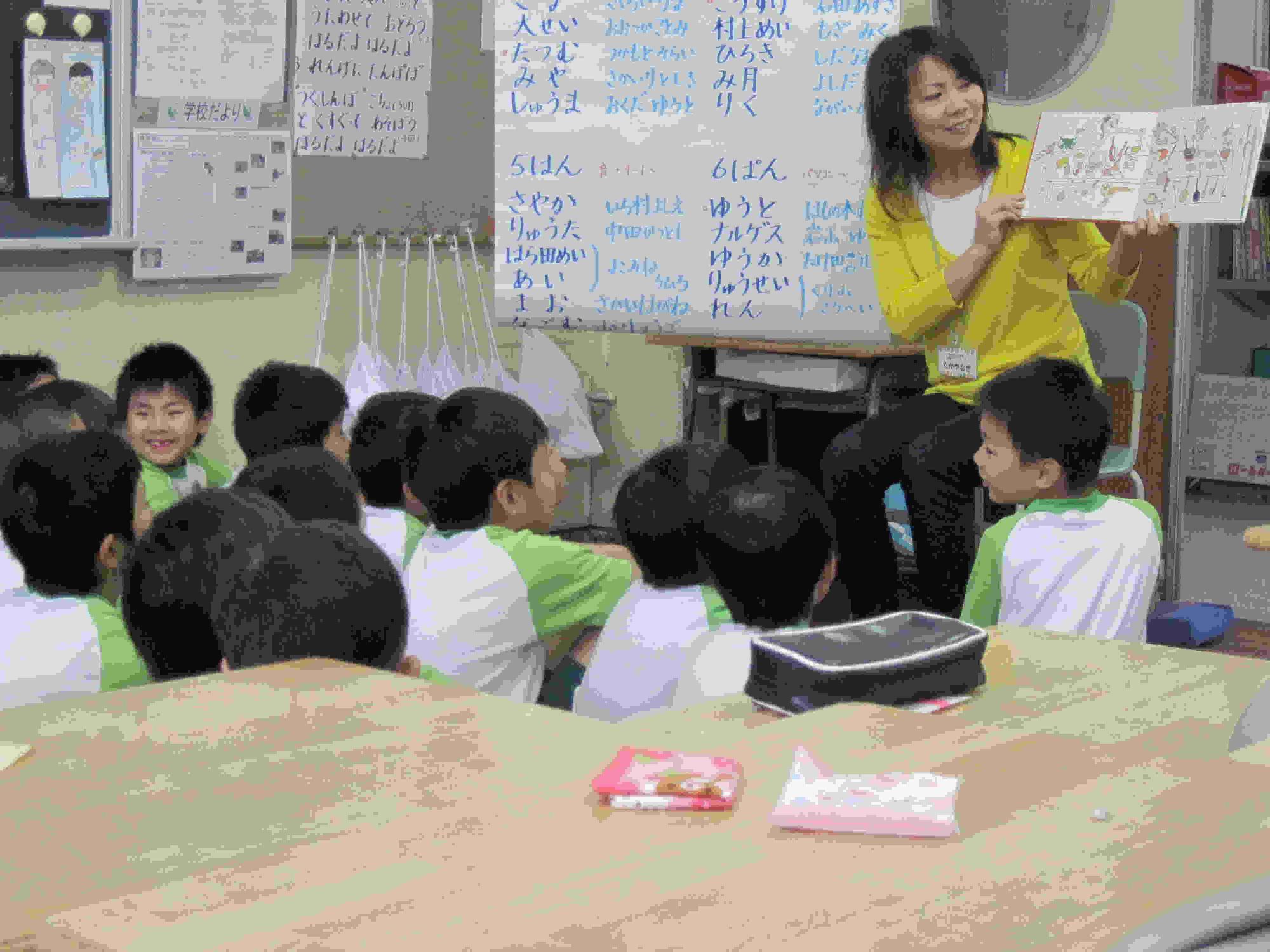 教室内前方で、椅子に座り絵本を開きながら、読み聞かせをするボランティアの方と、笑顔で読み聞かせを聞く児童たちの写真