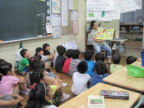 教室内で、椅子に座り絵本を開いて読み聞かせをするボランティアの方と、体育座りをしながら、大人しく読み聞かせを聞いている児童たちの写真