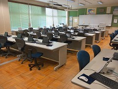緑のブラインドが設置され、白く細長いオフィスデスクの上にノートパソコンがいくつも並んでいる様子のパソコン室の写真