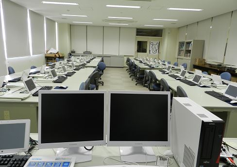 デュアルディスプレイのパソコンの机と、その奥に生徒向けのタブレットパソコンが並べられている写真